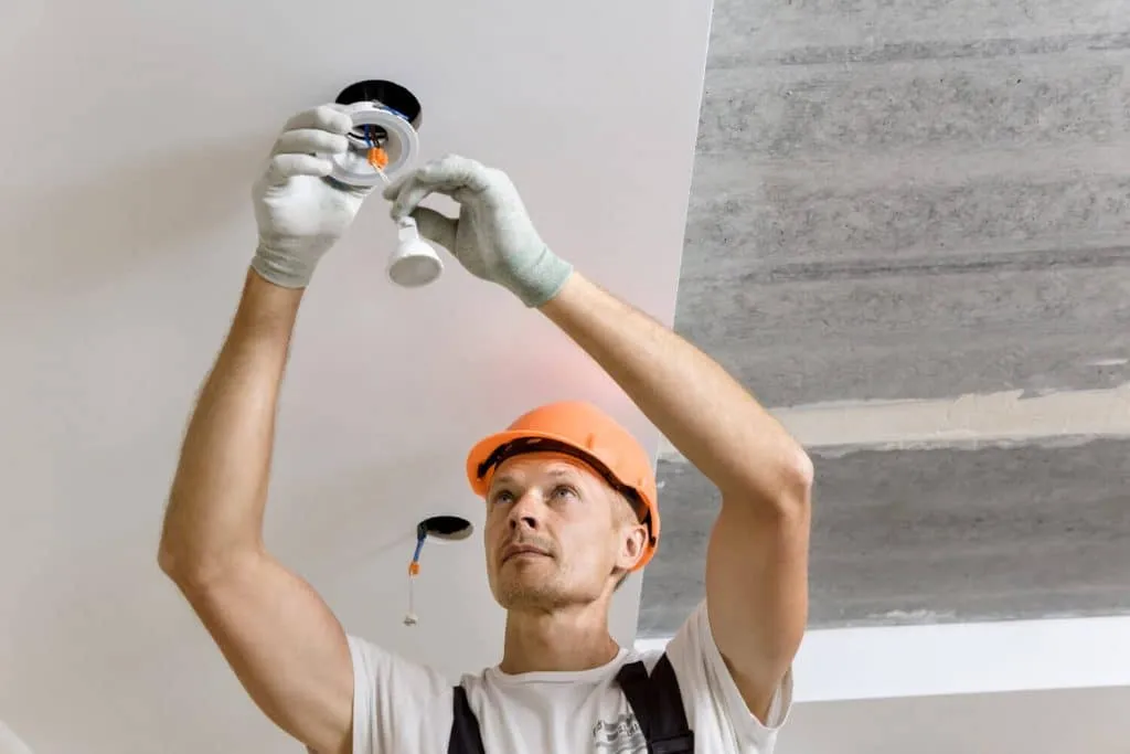 Electrician wearing a hard hat installing a light fixture on the ceiling.