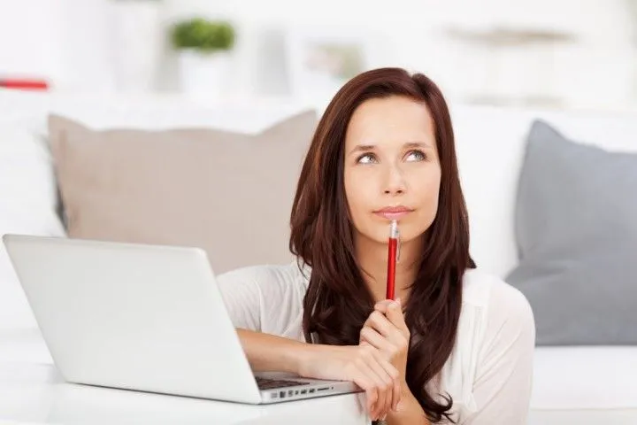Woman sitting with a laptop, holding a pen to her chin while thinking.