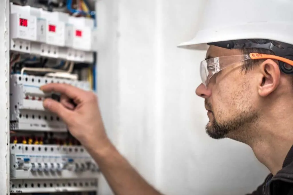 Electrician inspecting a breaker panel wearing safety glasses and a hard hat.