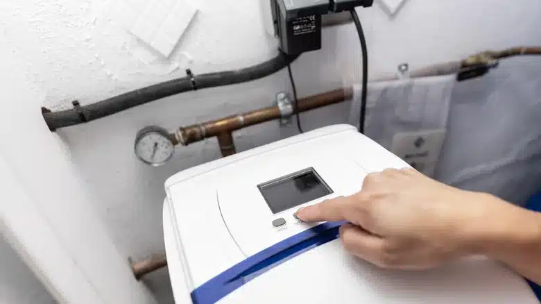 Person adjusting settings on a home water softener system with connected pipes and pressure gauge.